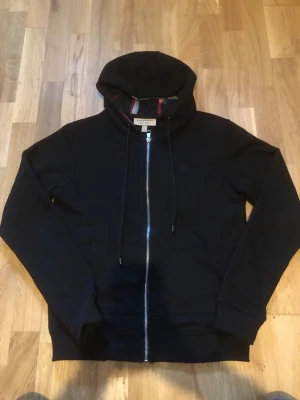 Svart hoodie med dragkedja från Burberry - Svart hoodie med dragkedja från Burberry. Klassisk huva med justerbara snören och ikoniskt rutigt foder i huvan och längs insidan. Mjukt material och ribbade muddar vid ärmslut och nederkant. Perfekt för en stilren och avslappnad look.