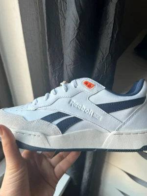 Vita Reebok sneakers med blå detaljer - Snygga vita Reebok sneakers med mörkblå detaljer och orange öljett. Klassisk låg modell med Reebok-logga på sidan. Ovandel i mocka och textil, sula i gummi. Perfekta för en sportig och clean stil.
