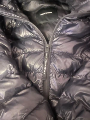 Svart dunjacka från Peak Performance - Snygg svart dunjacka från Peak Performance med glansig finish och diskret logga på bröstet. Jackan har dragkedja framtill och quiltad design som håller värmen under kalla dagar. Perfekt för dig som vill ha både stil och funktion i vinter nåra små hål på armarna inget som syns men därför ja säljer billigt