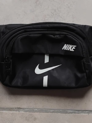 Svart midjeväska från Nike - Snygg svart midjeväska från Nike med vit logga och dragkedjor både fram och bak. Väskan är tillverkad i slitstarkt syntetmaterial och har flera fack för smart förvaring ( 4 stycken ). Perfekt accessoar för att hålla koll på dina grejer när du är på språng. Hör av er vid några frågor!