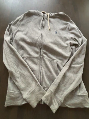 Grå zip hoodie från Polo Ralph Lauren - Snygg grå hoodie med dragkedja från Polo Ralph Lauren. Klassisk design med huva och vita snören, känguruficka framtill och den ikoniska lilla loggan broderad på bröstet. Tillverkad i mjukt bomullsmaterial som är perfekt för chill dagar.