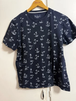 Mörkblå t-shirt med palmtryck - Snygg mörkblå t-shirt från Brooksfield med vitt palm- och ö-tryck över hela plagget. Klassisk rund halsringning och korta ärmar. Tillverkad i mjuk bomull som känns skön mot huden. Perfekt för dig som gillar en avslappnad och trendig stil.