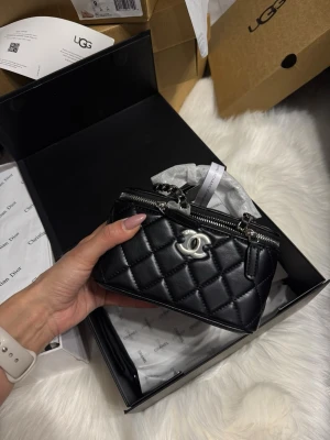 Svart quiltad handväska  - Säljer en svart quiltad handväska  med silverfärgad logga och dragkedja. Väskan har en boxig form, kedjerem och spegel på insidan av locket. Tillverkad i glansigt läder som ger en lyxig känsla. Perfekt för dig som gillar klassisk och trendig design.