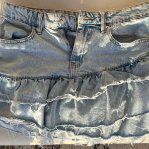 Lågmidjad Jeanskjol med volanger - Snygg ljusblå jeanskjol från Gina med volangdetaljer och råa, fransiga kanter. Jag säljer då den inte har kommit till användning. Den är som ny.