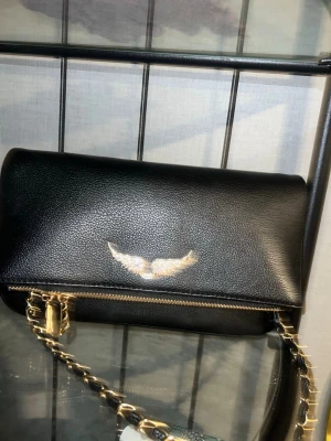 Svart handväska med guldkedja - Snygg svart handväska i läderimitation med guldfärgad kedjerem och ett broderat guldigt vingmotiv på framsidan. Väskan har ett stilrent och modernt utseende med dragkedja upptill. Perfekt accessoar för att lyfta din outfit. Använd bara några gånnger bara men kommer ej till användning 