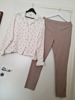 Kappahl Söt pyjamas med hjärtan och rutiga byxor - Kappahl mysig pyjamas med en vit skjorta med små bruna hjärtan och knappar framtill. Tillhörande byxor i beige med diskret rutmönster. Skjortan har krage och långa ärmar med volangdetalj vid ärmslut. Perfekt för chillkvällar hemma. Storlek L. Använd en gång, som ny.