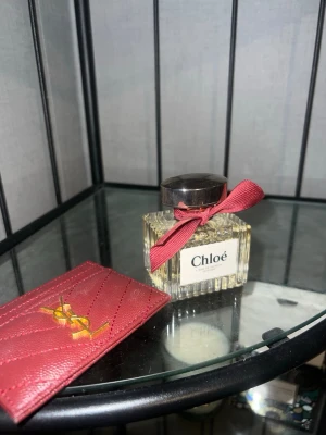 Chloé Eau de Parfum med röd rosett - Chloé Eau de Parfum i en elegant, fyrkantig glasflaska med räfflad design och en mörk metallkork. Flaskan är dekorerad med en röd rosett runt halsen som ger en söt touch. Perfekt för dig som gillar klassiska och feminina dofter.