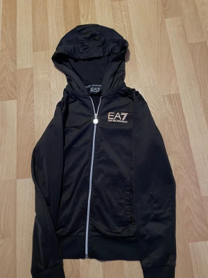 Svart hoodie från EA7 Emporio Armani - Snygg svart hoodie från EA7 Emporio Armani med dragkedja framtill och huva. Loggan i guld syns tydligt på bröstet och dragkedjan har en rund metalltag. Perfekt för en stilren och sportig look.