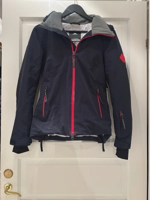 Skidjacka J.Lindeberg - Snygg marinblå skidjacka från J.Lindeberg med röda dragkedjor och detaljer. Älskar detta skisser så troligt mycket men måste sälja då det blivit för litet.  Jackan har en luva som går att dra över skihjälmen och även dragkedja på sidorna för varma dagar. Nypris för jacka: 4000kr, Byxor: 3000kr. Säljs dessvärre inte längre. Kan sälja för jacka: 3000kr då den ser helt ny ut. Byxor: 1500 då det är lite slitage vid rumpan. Eller hela setet för 4000kr💕Båda är i storlek S.