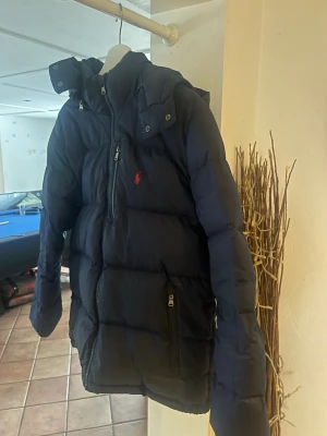 Mörkblå pufferjacka från Polo Ralph Lauren - Snygg mörkblå pufferjacka från Polo Ralph Lauren med huva och dragkedja framtill. Jackan har en röd logga på bröstet, två sidofickor med dragkedja och quiltad design. Perfekt för kalla vinterdagar och riktigt varm.