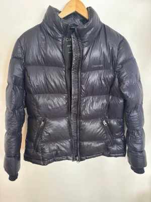 Svart pufferjacka från Calvin Klein - Snygg svart pufferjacka från Calvin Klein med hög krage och två dragkedjeförsedda fickor. Jackan har en glansig finish och quiltad design som ger en modern look. Perfekt för kalla dagar och enkel att matcha med olika outfits.