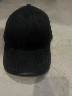 Svart keps från Gucci med GG-mönster - Snygg svart keps från Gucci med det klassiska GG-mönstret över hela kepsen. Tillverkad i mesh-liknande material med justerbar rem bak i läder och diskret Gucci-logga. Perfekt för dig som vill ha en stilren och exklusiv accessoar.