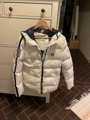 Vit pufferjacka från Dolomite - Säljer en snygg vit pufferjacka från Dolomite med marinblå detaljer och huva. Jackan har dragkedja framtill, två stora fickor och en patch på ärmen. Insidan har italienska flaggans färger och märkeslogga på bröstet. Perfekt för kalla vinterdagar.