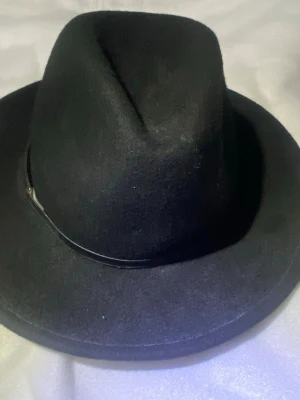 Svart fedora från Calvin Klein - Stilren svart fedora-hatt från Calvin Klein med klassisk form och smalt band runt kullen. Hatten är tillverkad i filtmaterial och har en diskret metallogo på bandet. Perfekt accessoar för att lyfta din outfit.