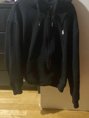 Svart hoodie från Polo Ralph Lauren - Svart zip-hoodie från Polo Ralph Lauren med vit broderad logga på bröstet. Tröjan har huva med dragsko, fickor framtill och ribbade muddar. Perfekt för en avslappnad och stilren look.