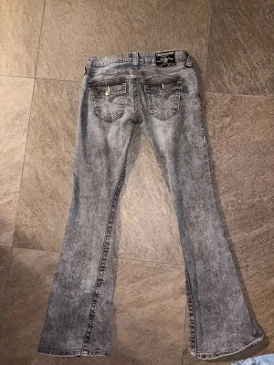 Grå bootcut jeans från True Religion Lågmidjade Joey - Säljer ett par grå tvättade bootcut jeans från True Religion med snygga detaljer på bakfickorna och klassisk logga på linningen. Jeansen har en lätt utsvängd passform och cool vintagekänsla. Perfekta för dig som gillar street och trendiga looks.