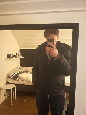 Svart pufferjacka från Ralph Lauren - Snygg svart pufferjacka från Ralph Lauren med röd logga på bröstet. Jackan har dragkedja framtill, hög krage och praktiska fickor. Perfekt för kalla dagar och har en klassisk, stilren look som passar till det mesta.