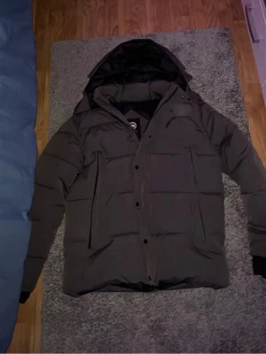 Svart dunjacka från Canada Goose - Säljer en svart dunjacka från Canada Goose med huva och klassisk patch på ärmen. Jackan har dragkedja och knappar framtill samt två stora fickor. Perfekt för kalla dagar och riktigt varm med sin vaddering.