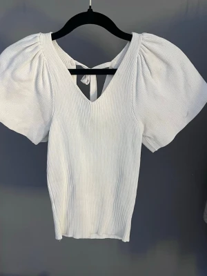 Vit ribbad topp med puffärm och knyt - Supersnygg vit ribbad topp med puffiga korta ärmar och v-ringning både fram och bak. Toppen har ett gulligt knyt i ryggen som detalj och är i ett mjukt, stretchigt material. Perfekt för dig som vill ha en trendig och bekväm look.