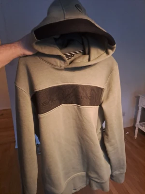 Olivgrön Sail Racing hoodie - Snygg olivgrön hoodie från Sail Racing med svart panel och diskret logga på bröstet. Hoodien har en stor huva och ribbade muddar vid ärmar och nederkant. Tillverkad i mjukt bomullsmaterial som känns skönt mot huden. Perfekt för en avslappnad streetwear-look.