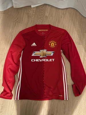 Manchester United långärmad matchtröja - Röd långärmad Manchester United matchtröja från Adidas med klubbmärke och Chevrolet-logga på bröstet. Tröjan har vita ränder längs sidorna och är tillverkad i lätt funktionsmaterial (climacool). Perfekt för fotbollsträning eller att bära på läktaren.