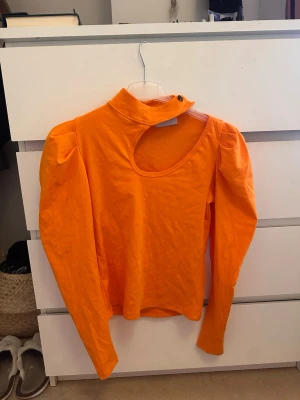 Orange långärmad topp med cut out - Trendig orange långärmad topp med unik cut out-detalj vid halsen och axelparti. Puffiga axlar och hög krage med knappstängning ger en cool och modern look. Perfekt för dig som vill sticka ut med färg och form.