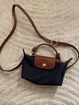 Mörkblå axelväska från Longchamp - Snygg mörkblå axelväska från Longchamp med justerbar brun axelrem och detaljer i brunt läder. Väskan har guldfärgade metalldetaljer och klassiskt präglat logotyplock. Perfekt storlek för det viktigaste och enkel att matcha till olika outfits.