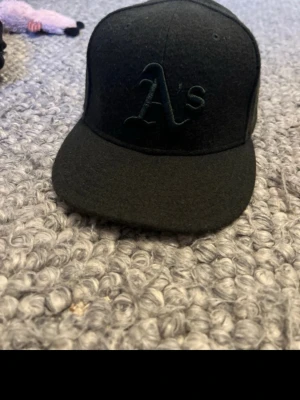 Svart New Era Oakland A's keps - Snygg svart keps från New Era med Oakland A's-logga broderad framtill i samma färg som kepsen. Klassisk flat brim och strukturerad form. Perfekt för dig som gillar sportig streetstyle och vill ha en stilren look.