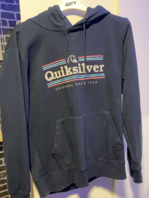 Mörkblå Quiksilver hoodie - Snygg mörkblå hoodie från Quiksilver med stor logga och texten 'Original Since 1969' på bröstet. Klassisk känguruficka framtill, huva med dragsko och ribbade muddar. Perfekt för en avslappnad och cool stil.