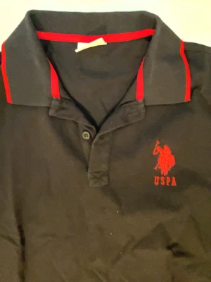 Svart USPA pikétröja med röda detaljer - Svart pikétröja från USPA med röda detaljer på krage och ärmslut samt röd logga på bröstet. Klassisk pikékrage med knappslå framtill. Perfekt för en sportig och stilren look.