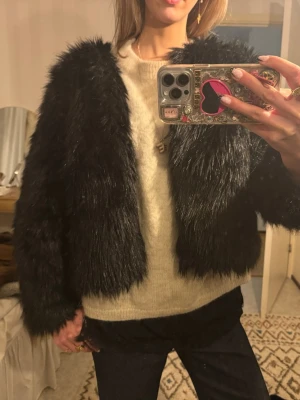 Svart fuskpälsjacka - Snygg svart fuskpälsjacka i kort modell med lång ärm och rundad hals. Jackan har ett fluffigt och glansigt pälsmaterial som ger en lyxig känsla. Perfekt för att höja din outfit under kalla dagar.
