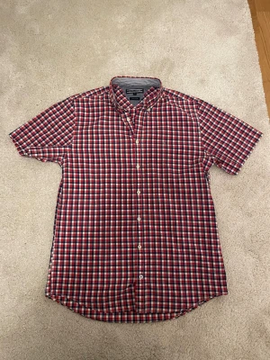Rutig kortärmad skjorta Tommy Hilfiger - Snygg kortärmad skjorta från Tommy Hilfiger i rött, blått och vitt rutigt mönster. Klassisk button-down krage och vita knappar framtill. Perfekt för dig som gillar en avslappnad men ändå stilren look.