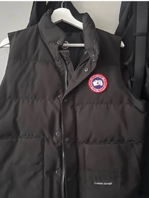 Svart dunväst från Canada Goose - Snygg svart dunväst från Canada Goose med klassisk logopatch på bröstet och två stora fickor framtill. Västen har hög krage, quiltad design och dragkedja med tryckknappar. Perfekt för lager-på-lager under kyliga dagar. storlek 180cm