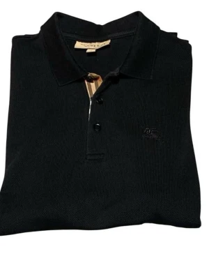 Svart pikétröja från Burberry - Klassisk svart pikétröja från Burberry med ikonisk beige-rutig detalj vid knäppningen och diskret broderad logga på bröstet. Tröjan har krage, korta ärmar och knappar framtill. Perfekt för en stilren och tidlös look.