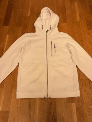 Vit  Zip Hoodie från Sail Racing - Snygg vit hoodie från Sail Racing med dragkedja och klassisk logga broderad på bröstet. Tröjan har huva med snörning och två stora fickor framtill, samt en ficka med dragkedja vid bröstet. Perfekt för en clean och stilren look.                                           
