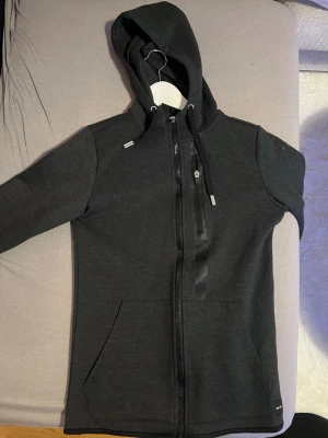 Svart hoodie med dragkedja - Mörkgrå hoodie med hel dragkedja framtill, huva med dragsko och en bröstficka med dragkedja. Två sidofickor och diskreta detaljer i svart. Perfekt för chill dagar eller när du vill ha en clean look.