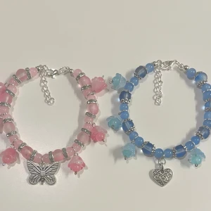 Två armband med berlocker - HANDGJORT Två armband med pärlor, ett i rosa och ett i blått. Båda har små blomformade pärlor och silverfärgade detaljer. Det rosa armbandet har en fjärilsberlock och det blå ett hjärta. Det finns en en förlängnings kedja.
