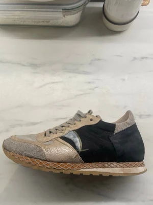 Philippe Model beige och svarta sneakers - Snygga sneakers från Philippe Model i beige och svart med metallicdetaljer och ormskinnsmönstrad textur. Skon har en klassisk låg silhuett, robust gummisula och coola kontrastpaneler. Perfekt för dig som vill sticka ut med stilrena detaljer.