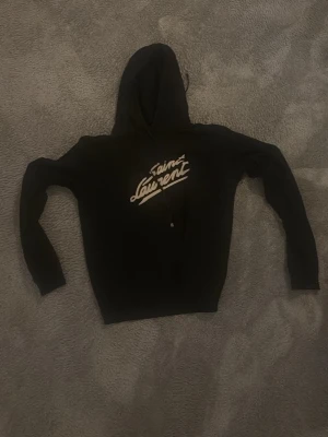 Svart Saint Laurent hoodie med tryck - Svart hoodie från Saint Laurent med vit logotyp på bröstet i snyggt tryck. Klassisk modell med huva och dragsko. Perfekt för dig som gillar streetwear och vill ha något stilrent men ändå trendigt. Mjuk och skön bomullskänsla.