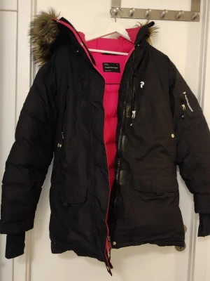 Svart Peak Performance dunjacka - Säljer en svart dunjacka från Peak Performance med rosa innerfoder och avtagbar pälskant på huvan. Jackan har flera fickor med dragkedja, justerbara muddar och en snygg, sportig look. Perfekt för kalla vinterdagar.