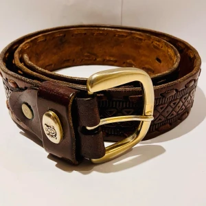 Brunt skärp i skinn med mönster - Snyggt brunt bälte i skinn med ingraverat geometriskt mönster och rejält mässingsfärgat spänne. Perfekt accessoar för att lyfta din outfit med en klassisk touch. Bältet har detaljerade nitar och präglad design längs hela remmen.