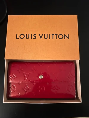 Röd Louis Vuitton plånbok lack - Snygg röd plånbok från Louis Vuitton i glansigt lackat läder med klassiskt monogrammönster. Plånboken har en knappstängning i metall och kommer i originalask. Perfekt accessoar för dig som gillar lyxiga detaljer och ikonisk design.