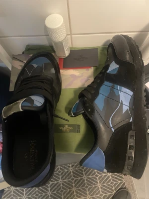Valentino roxkrunners - Säljer ett par snygga sneakers från Valentino med blått och grått camo-mönster. Skorna har svarta detaljer, svarta skosnören och coola nitar på sulan. Tillverkade i en mix av läder och textil för en exklusiv känsla.endast använda ett fåtal gånger, Då det inte är min stil direkt