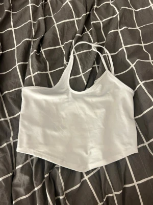 Vit ärmlös bandeau topp - Säljer en vit bandeau topp med halterneck och smala axelband. Toppen är croppad och har en enkel, stilren design som passar perfekt till sommaren. Materialet känns mjukt och stretchigt, troligen polyester eller bomull.