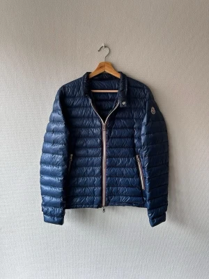 Mörkblå dunjacka från Moncler - Snygg mörkblå dunjacka från Moncler med quiltad design och två dragkedjeförsedda fickor. Jackan har en klassisk Moncler Daniel, perfekt för lite kallare vårdagar. Köpt från Merchsweden kvitto finns. Finns ett litet märke inget man tänker på annars fint skick, hör av er vid minsta fundering