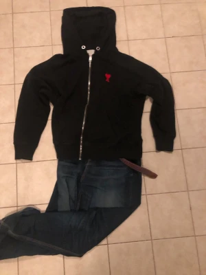 Svart hoodie med röd Ami-dekor - Fet tröja från AMI Paris med dragkedja framtill och huva till ett riktigt bra pris . Pris går att diskutera. Om du har några funderingar så är det bara att säga till!                                   OBS Storlek M men sitter som S