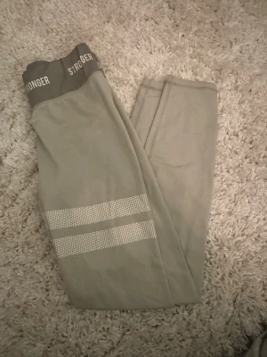 Beige leggings från Stronger - Snygga beige/grön leggings från Stronger med bred resår i midjan där loggan STRONGER syns. Perfekta för träning eller chill. Mjukt och stretchigt material för skön passform.