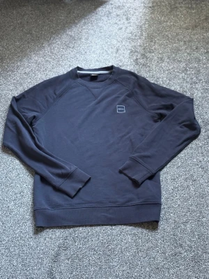 Mörkblå sweatshirt från BOSS - Säljer en stilren mörkblå sweatshirt från BOSS med rund halsringning och diskret logotyp på bröstet. Tröjan har långa ärmar, ribbade muddar och är tillverkad i mjukt bomullsmaterial. Perfekt för en clean och avslappnad look.