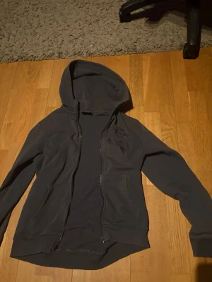 Mörkgrå hoodie Peak Performance - Snygg mörkgrå hoodie från Peak Performance med dragkedja framtill och broderad logga på bröstet. Hoodien har två fickor fram och en klassisk huva. Tillverkad i mjukt bomullsmaterial som känns skönt mot huden. Perfekt för en avslappnad och sportig stil.