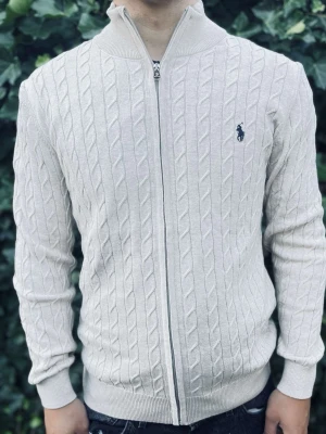 Vit kabelstickad ziptröja Polo Ralph Lauren - Kabelstickad vit ziptröja från Polo Ralph Lauren med broderad logga på bröstet. Tröjan har hög krage, hel dragkedja och ribbade muddar. Perfekt för lager-på-lager och snygg till jeans.